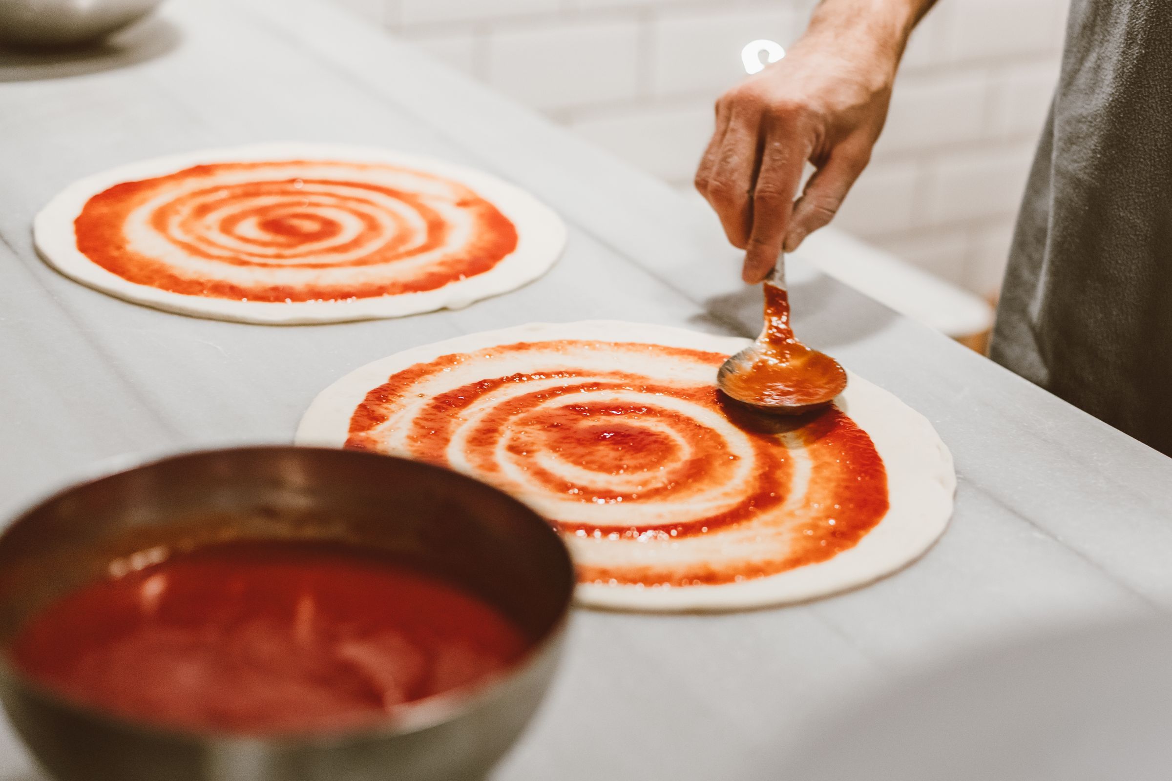 Tomato sauce being spread over pizza dough with a spoon
